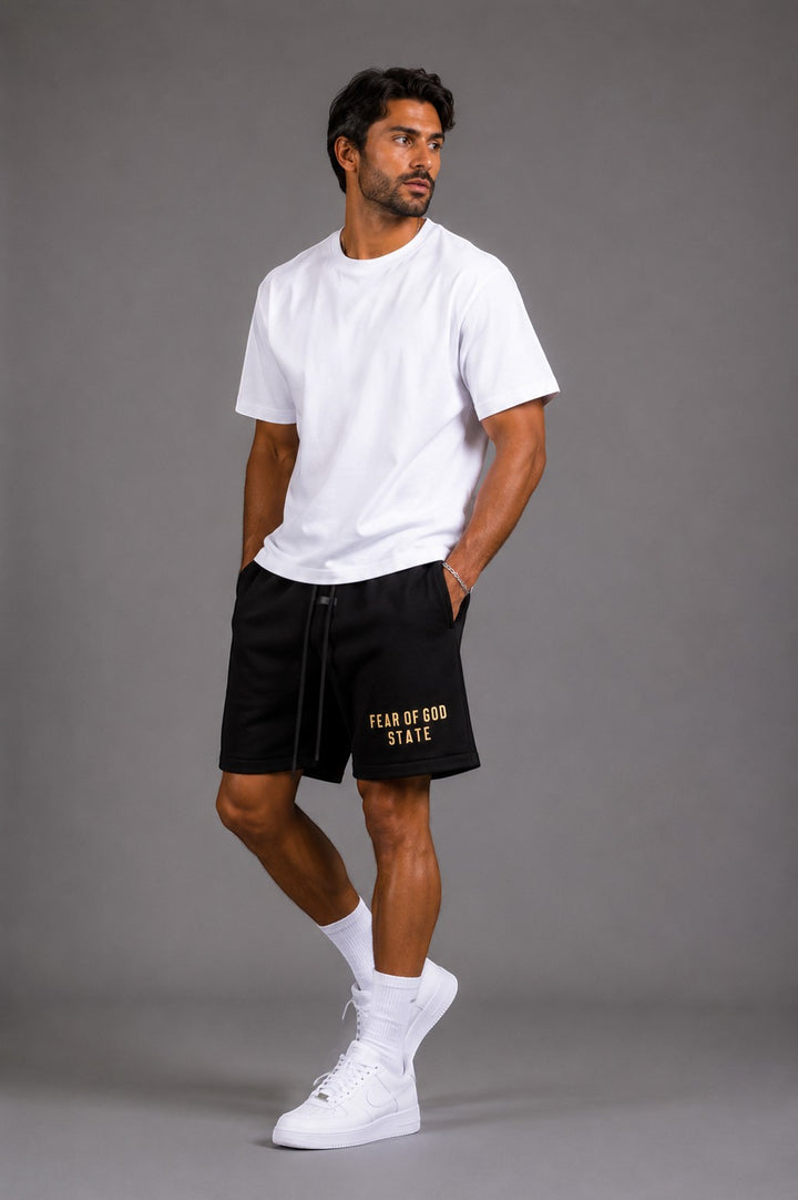 Hoopers black Fear Of God Shadow State shorts with gold logo, drawstring waist, and side pockets. Model wears white t-shirt and sneakers.